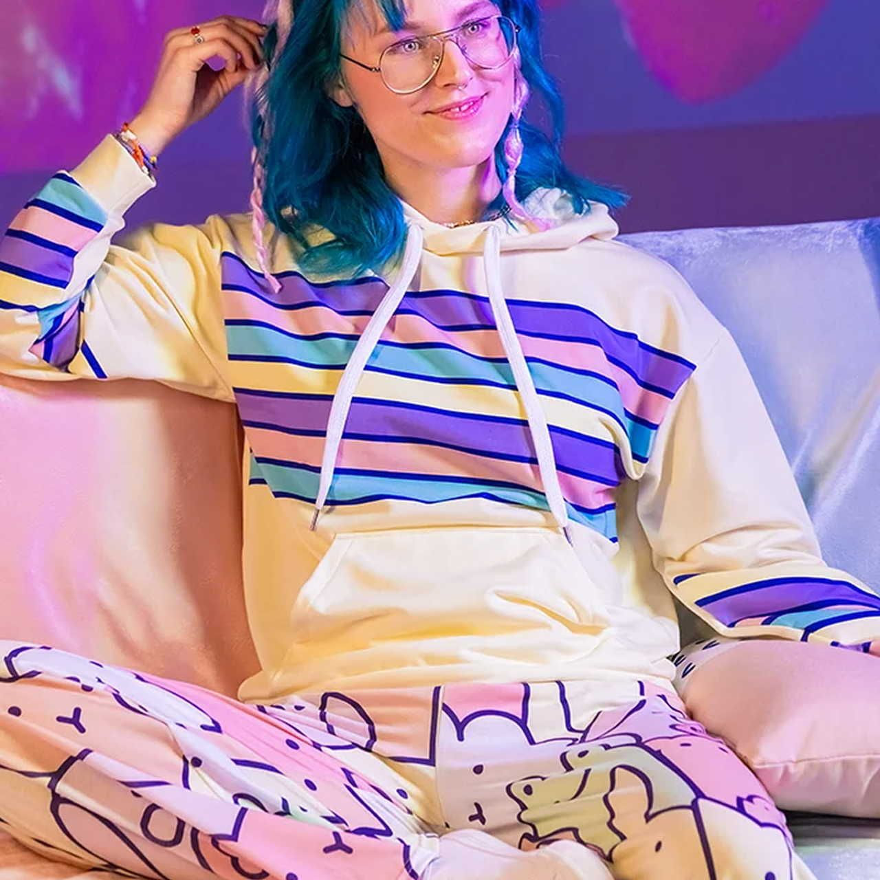 Woman with blue hair and glasses, wearing a pastel-striped hoodie, sits on a sofa with a relaxed smile. Her pants feature abstract patterns.